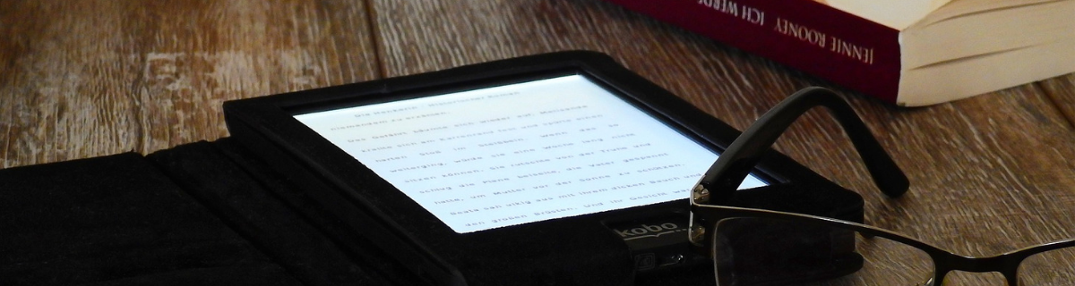 image: e-reader, paperback book, and reading glasses on brown wooden table